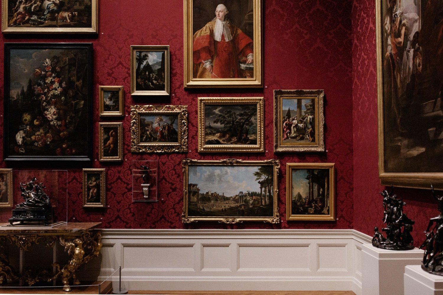 A salon-style hang of framed paintings on deep red damask
                     wallpaper above a white dado.