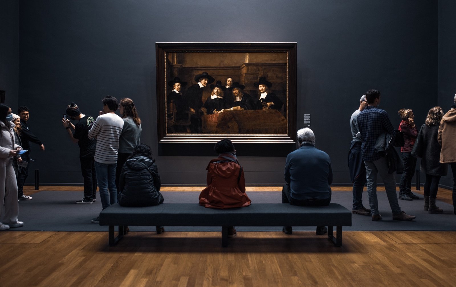 Synthesis — visitors seated on a bench before a large
                         group portrait in a dark gallery.
